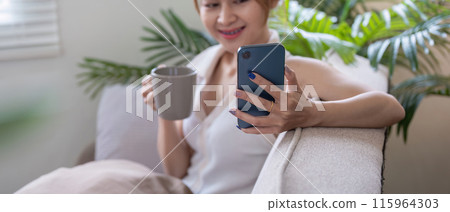 Woman Relaxing at Home Using Smartphone for Social Media While Drinking Coffee in a Cozy Living Room Woman Relaxing at Home Using Smartphone for Social Media While Drinking Coffee in a Cozy Living Room 115964303