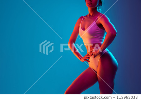 Young woman with toned smooth skin posing in lingerie hold hands on waist in pink neon light against gradient blue background. Young woman with toned smooth skin posing in lingerie hold hands on waist in pink neon light against gradient blue background. 115965038