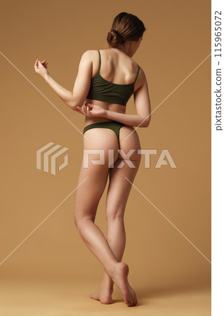 Rear view photo of young beautiful woman posing holds hand behind back showing her figure against sandy studio background. 115965072