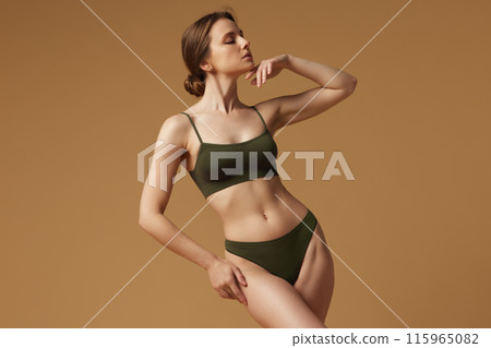 Slim woman posing in khaki lingerie gently raising hands demonstrating her sensuality and perfect figure against sandy studio background. Slim woman posing in khaki lingerie gently raising hands demonstrating her sensuality and perfect figure against sandy studio background. 115965082