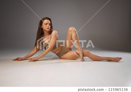 Emphasizing body care, woman's glowing skin and serene expression in beige underwear reflect her wellness regimen against warm grey studio background. 115965136