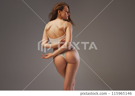 Rear view photo of woman poses in white lingerie, showcasing her healthy back against warm grey studio background. Slim model. Rear view photo of woman poses in white lingerie, showcasing her healthy back against warm grey studio background. Slim model. 115965146