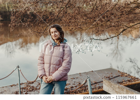 A young woman standing at the shore looking at the river in autumn sunny day. Street view, copy space for text, travel photo. Happy tourist woman on the bank of the river in autumn in warm clothes 115967661