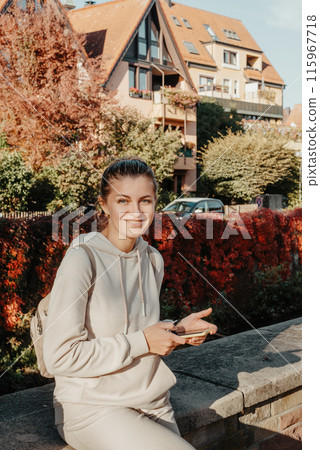 Young Fashionable Teenage Girl With Smartphone In Europian Park In Autumn Sitting At Smiling. Trendy Young Woman In Fall In Park Texting. Retouched, Vibrant Colors. Beautiful Blonde Teenage Girl 115967718
