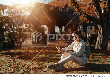 Young Fashionable Teenage Girl With Smartphone In Europian Park In Autumn Sitting At Smiling. Trendy Young Woman In Fall In Park Texting. Retouched, Vibrant Colors. Beautiful Blonde Teenage Girl 115967725