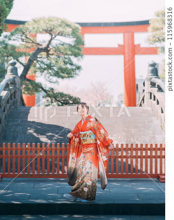 A woman in a long-sleeved kimono standing at the torii gate of a shrine 115968316