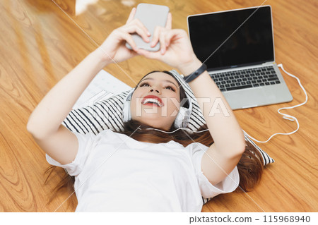 Top view of young beautiful asian woman with headphone listening music from laptop and using smartphone  while lying on the floor at home. 115968940