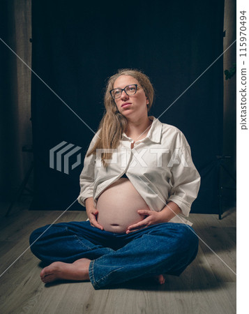 Portrait of a pregnant woman sitting cross-legged on the floor, white shirt unbuttoned, exposing her bare belly. Dramatic atmosphere of anticipation and the beauty of motherhood. 115970494
