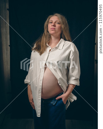 Portrait of a pregnant woman standing against a dark background. She is wearing a white unbuttoned shirt, exposing her belly, and denim trousers. Her left hand is slipped carelessly into her pocket. Portrait of a pregnant woman standing against a dark background. She is wearing a white unbuttoned shirt, exposing her belly, and denim trousers. Her left hand is slipped carelessly into her pocket. 115970495