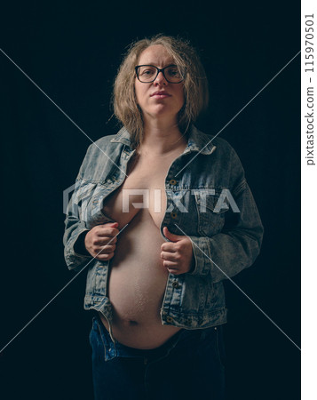 Belt portrait of pregnant woman wearing unbuttoned denim jacket and blue jeans on dark background. Exposing her bare torso and belly. The beauty of late term pregnancy. Belt portrait of pregnant woman wearing unbuttoned denim jacket and blue jeans on dark background. Exposing her bare torso and belly. The beauty of late term pregnancy. 115970501