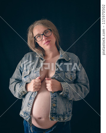 Portrait of a pregnant woman against a dark background. She is wearing a partially open denim jacket over her naked body. Concept of the joy of pregnancy. Portrait of a pregnant woman against a dark background. She is wearing a partially open denim jacket over her naked body. Concept of the joy of pregnancy. 115970502