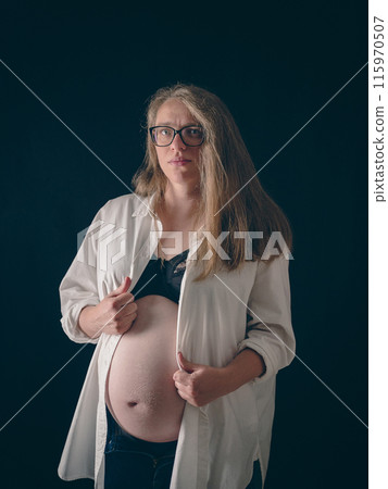 Pregnant woman wearing glasses and casual clothes partially unbuttoned white shirt and blue jeans on black background. She holds up her shirt with one hand, exposing her black bra. Belt portrait. Pregnant woman wearing glasses and casual clothes partially unbuttoned white shirt and blue jeans on black background. She holds up her shirt with one hand, exposing her black bra. Belt portrait. 115970507