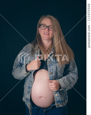 A mom-to-be in late pregnancy wearing an unbuttoned denim jacket. Belt portrait of a pregnant woman on a dark background. Joy and anticipation of motherhood. A mom-to-be in late pregnancy wearing an unbuttoned denim jacket. Belt portrait of a pregnant woman on a dark background. Joy and anticipation of motherhood. 115970509