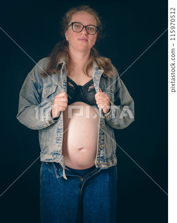 An expectant mother stands with her denim jacket open, revealing her pregnant belly. She wears a black top and blue jeans, embodying the anticipation and beauty of motherhood. 115970512