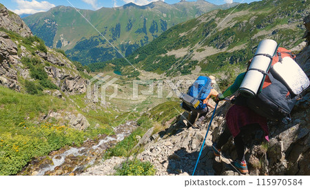 Mountain Adventure. A group of hikers with large rucksacks descend a steep mountain path in high altitude conditions, using trekking poles. A stream flows nearby. Mountain Adventure. A group of hikers with large rucksacks descend a steep mountain path in high altitude conditions, using trekking poles. A stream flows nearby. 115970584