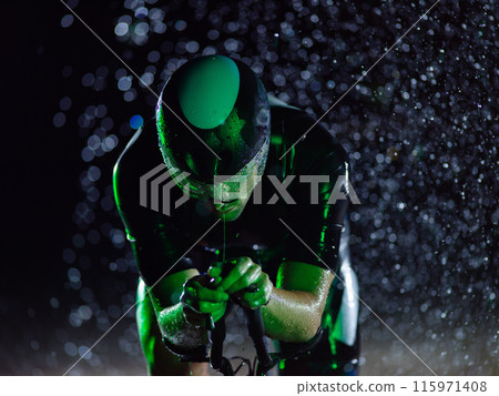 A triathlete braving the rain as he cycles through the night, preparing himself for the upcoming marathon. The blurred raindrops in the foreground and the dark, moody atmosphere in the background add 115971408