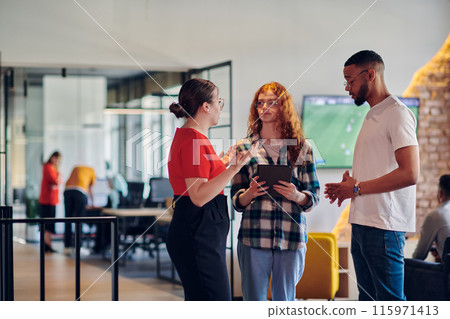 A group of young business individuals, including a girl with orange hair and an African American man, stands in a modern corporate hallway, collectively examining business progress on a smartphone 115971413
