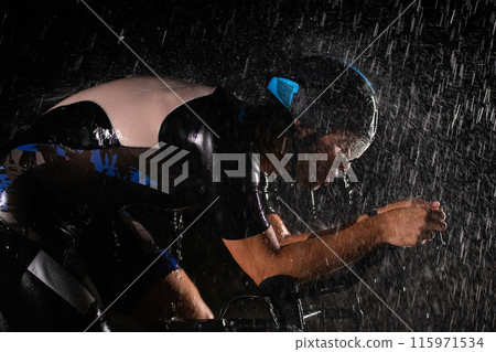 A triathlete braving the rain as he cycles through the night, preparing himself for the upcoming marathon. The blurred raindrops in the foreground and the dark, moody atmosphere in the background add 115971534