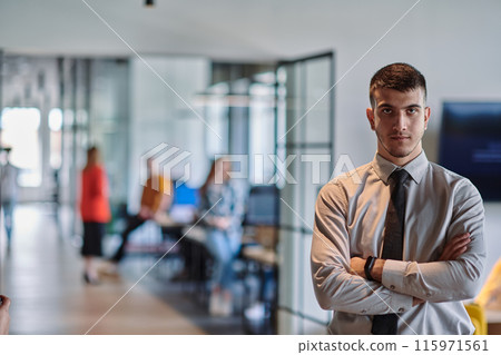 A young business leader stands with crossed arms in a modern office hallway, radiating confidence and a sense of purpose, embodying a dynamic and inspirational presence. 115971561