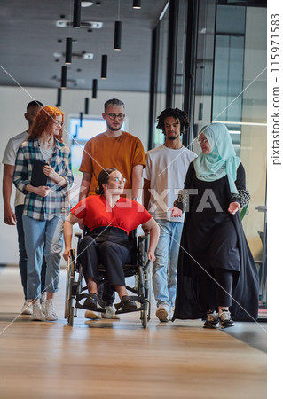 A diverse group of young business people walking a corridor in the glass-enclosed office of a modern startup, including a person in a wheelchair and a woman wearing a hijab, showing a dynamic mix of 115971583