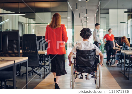 A group of young business people in a modern glass-walled office captures the essence of diversity and collaboration, while two colleagues, including an African American businessman in a wheelchair 115971586