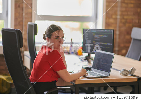 In a contemporary office setting, a young businesswoman is focused on her laptop, displaying dedication and efficiency in her work 115971699