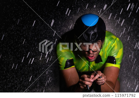 A triathlete braving the rain as he cycles through the night, preparing himself for the upcoming marathon. The blurred raindrops in the foreground and the dark, moody atmosphere in the background add 115971792