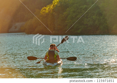 A young couple enjoying an idyllic kayak ride in the middle of a beautiful river surrounded by forest greenery in sunset time  115971851
