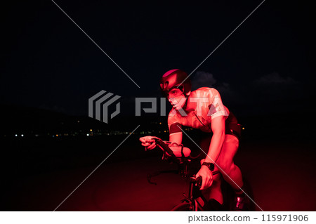 A triathlete rides his bike in the darkness of night, pushing himself to prepare for a marathon. The contrast between the darkness and the light of his bike creates a sense of drama and highlights the 115971906