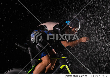 A triathlete braving the rain as he cycles through the night, preparing himself for the upcoming marathon. The blurred raindrops in the foreground and the dark, moody atmosphere in the background add A triathlete braving the rain as he cycles through the night, preparing himself for the upcoming marathon. The blurred raindrops in the foreground and the dark, moody atmosphere in the background add 115971907