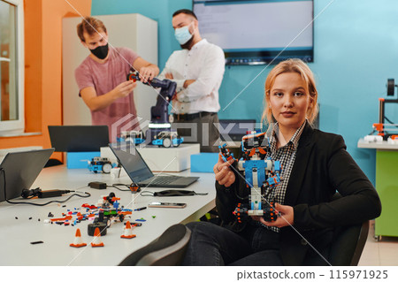 A woman sitting in a laboratory and solving problems and analyzing the robot's verification. In the background, colleagues are talking at an online meeting A woman sitting in a laboratory and solving problems and analyzing the robot's verification. In the background, colleagues are talking at an online meeting 115971925