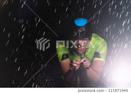 A triathlete braving the rain as he cycles through the night, preparing himself for the upcoming marathon. The blurred raindrops in the foreground and the dark, moody atmosphere in the background add 115971949