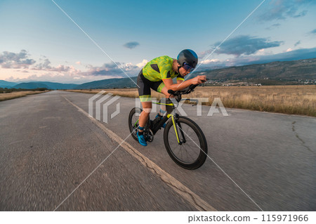 Triathlete riding his bicycle during sunset, preparing for a marathon. The warm colors of the sky provide a beautiful backdrop for his determined and focused effort. 115971966