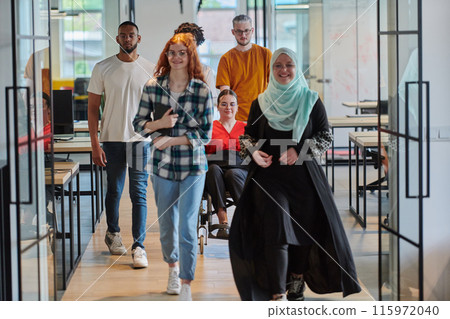 A diverse group of business people walking a corridor in the glass-enclosed office of a modern startup, including a person in a wheelchair and a woman wearing a hijab, showing a dynamic mix of 115972040