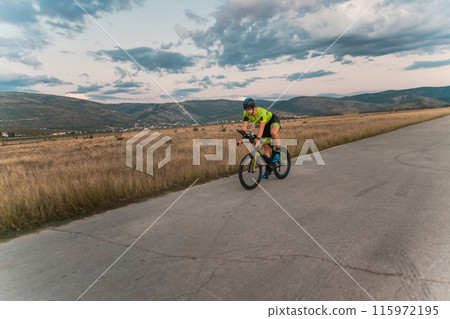 Triathlete riding his bicycle during sunset, preparing for a marathon. The warm colors of the sky provide a beautiful backdrop for his determined and focused effort. 115972195