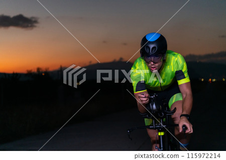 A triathlete rides his bike in the darkness of night, pushing himself to prepare for a marathon. The contrast between the darkness and the light of his bike creates a sense of drama and highlights the A triathlete rides his bike in the darkness of night, pushing himself to prepare for a marathon. The contrast between the darkness and the light of his bike creates a sense of drama and highlights the 115972214