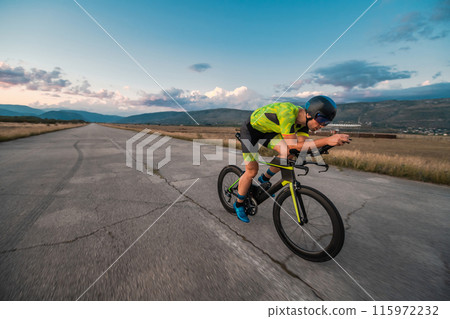 Triathlete riding his bicycle during sunset, preparing for a marathon. The warm colors of the sky provide a beautiful backdrop for his determined and focused effort. Triathlete riding his bicycle during sunset, preparing for a marathon. The warm colors of the sky provide a beautiful backdrop for his determined and focused effort. 115972232
