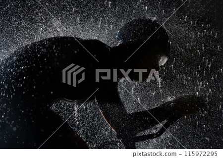 A triathlete braving the rain as he cycles through the night, preparing himself for the upcoming marathon. The blurred raindrops in the foreground and the dark, moody atmosphere in the background add 115972295