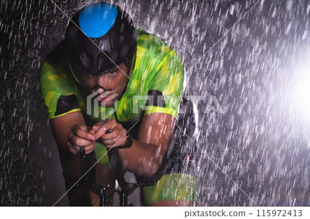 A triathlete braving the rain as he cycles through the night, preparing himself for the upcoming marathon. The blurred raindrops in the foreground and the dark, moody atmosphere in the background add A triathlete braving the rain as he cycles through the night, preparing himself for the upcoming marathon. The blurred raindrops in the foreground and the dark, moody atmosphere in the background add 115972413