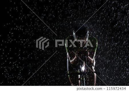 A triathlete braving the rain as he cycles through the night, preparing himself for the upcoming marathon. The blurred raindrops in the foreground and the dark, moody atmosphere in the background add A triathlete braving the rain as he cycles through the night, preparing himself for the upcoming marathon. The blurred raindrops in the foreground and the dark, moody atmosphere in the background add 115972434