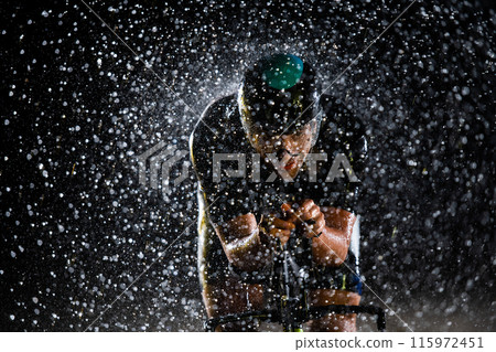 A triathlete braving the rain as he cycles through the night, preparing himself for the upcoming marathon. The blurred raindrops in the foreground and the dark, moody atmosphere in the background add A triathlete braving the rain as he cycles through the night, preparing himself for the upcoming marathon. The blurred raindrops in the foreground and the dark, moody atmosphere in the background add 115972451