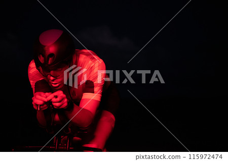 A triathlete rides his bike in the darkness of night, pushing himself to prepare for a marathon. The contrast between the darkness and the light of his bike creates a sense of drama and highlights the A triathlete rides his bike in the darkness of night, pushing himself to prepare for a marathon. The contrast between the darkness and the light of his bike creates a sense of drama and highlights the 115972474