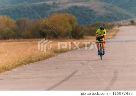 Triathlete riding his bicycle during sunset, preparing for a marathon. The warm colors of the sky provide a beautiful backdrop for his determined and focused effort. Triathlete riding his bicycle during sunset, preparing for a marathon. The warm colors of the sky provide a beautiful backdrop for his determined and focused effort. 115972483