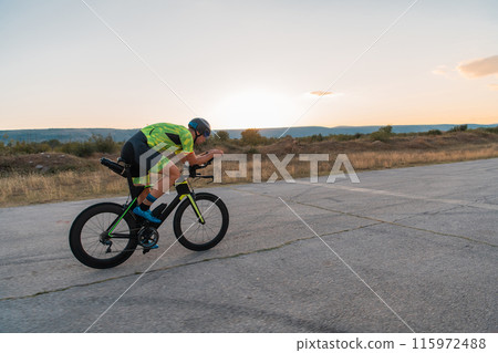 Triathlete riding his bicycle during sunset, preparing for a marathon. The warm colors of the sky provide a beautiful backdrop for his determined and focused effort. 115972488