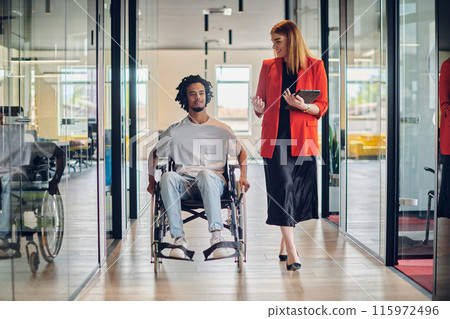 A group of young business people in a modern glass-walled office captures the essence of diversity and collaboration, while two colleagues, including an African American businessman in a wheelchair A group of young business people in a modern glass-walled office captures the essence of diversity and collaboration, while two colleagues, including an African American businessman in a wheelchair 115972496