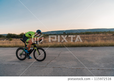 Triathlete riding his bicycle during sunset, preparing for a marathon. The warm colors of the sky provide a beautiful backdrop for his determined and focused effort. Triathlete riding his bicycle during sunset, preparing for a marathon. The warm colors of the sky provide a beautiful backdrop for his determined and focused effort. 115972516