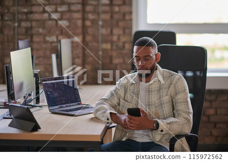A young African American entrepreneur takes a break in a modern office, using a smartphone to browse social media, capturing a moment of digital connectivity and relaxation amidst his business 115972562