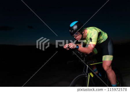 A triathlete rides his bike in the darkness of night, pushing himself to prepare for a marathon. The contrast between the darkness and the light of his bike creates a sense of drama and highlights the A triathlete rides his bike in the darkness of night, pushing himself to prepare for a marathon. The contrast between the darkness and the light of his bike creates a sense of drama and highlights the 115972578