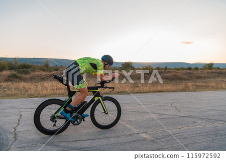 Triathlete riding his bicycle during sunset, preparing for a marathon. The warm colors of the sky provide a beautiful backdrop for his determined and focused effort. 115972592