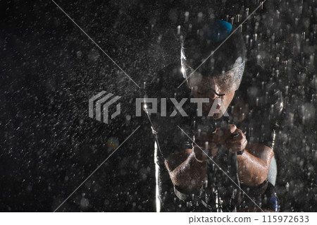 A triathlete braving the rain as he cycles through the night, preparing himself for the upcoming marathon. The blurred raindrops in the foreground and the dark, moody atmosphere in the background add A triathlete braving the rain as he cycles through the night, preparing himself for the upcoming marathon. The blurred raindrops in the foreground and the dark, moody atmosphere in the background add 115972633